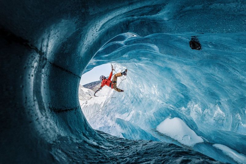 An ice climber hangs from an axe embedded in the blue walls of a circular ice shaft.