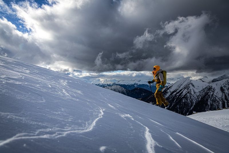 A climber ascends a snow-covered peak, mountains behind and dark skies overhead.