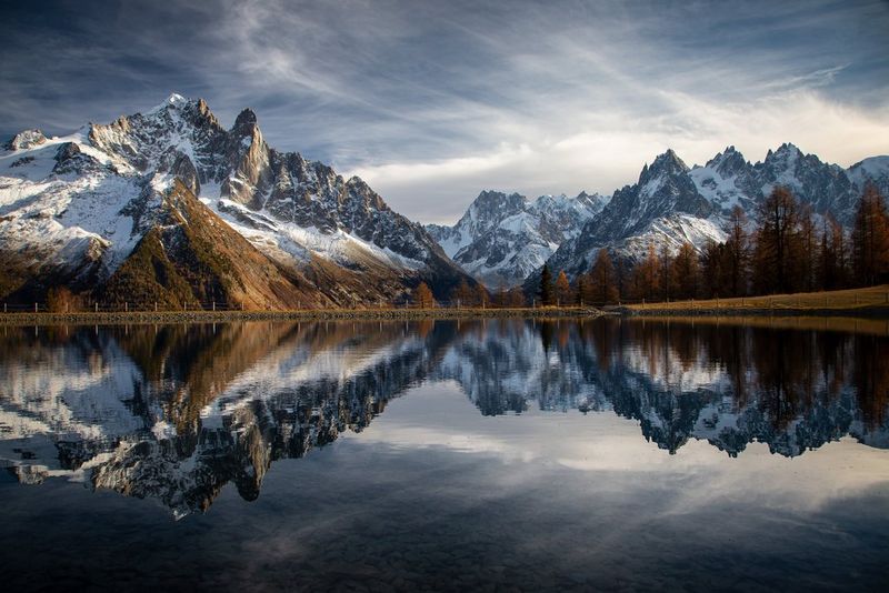 Snow-covered mountains rising up behind a line of orange-tinged trees reflected in the still waters of a lake. Taken by Canon Ambassador Julia Roger-Veyer. 
