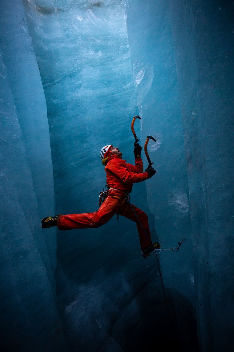 An ice climber in a red climbing suit hangs from two ice axes embedded in the blue walls of a vertical ice shaft, one leg on the wall behind him, the other in front. Taken by Canon Ambassador Julia Roger-Veyer. 