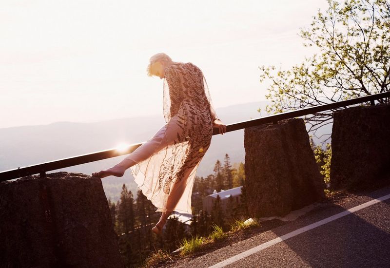 A model climbs on a roadside safety barrier in dappled sunshine. Below her is a sprawling green valley.