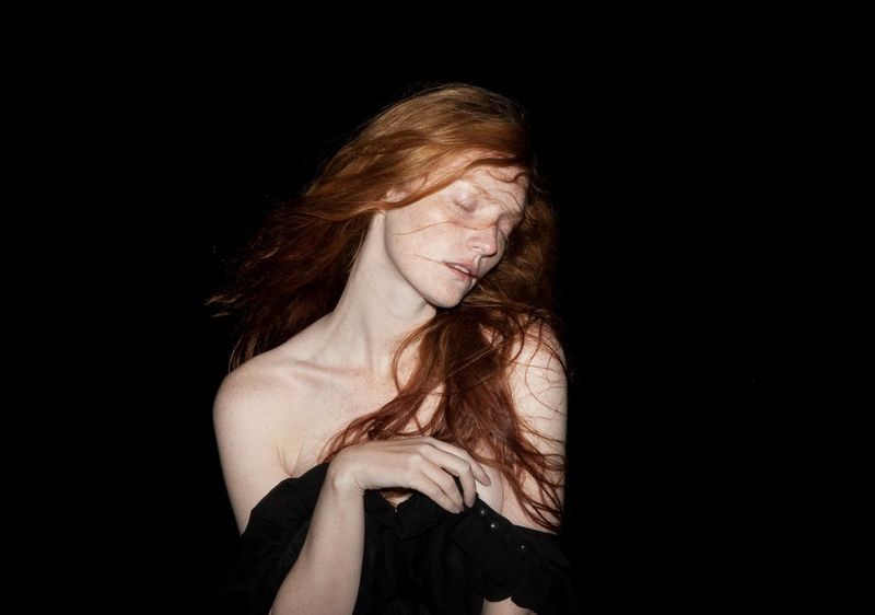 A red-headed model with her eyes closed is photographed against a black backdrop.