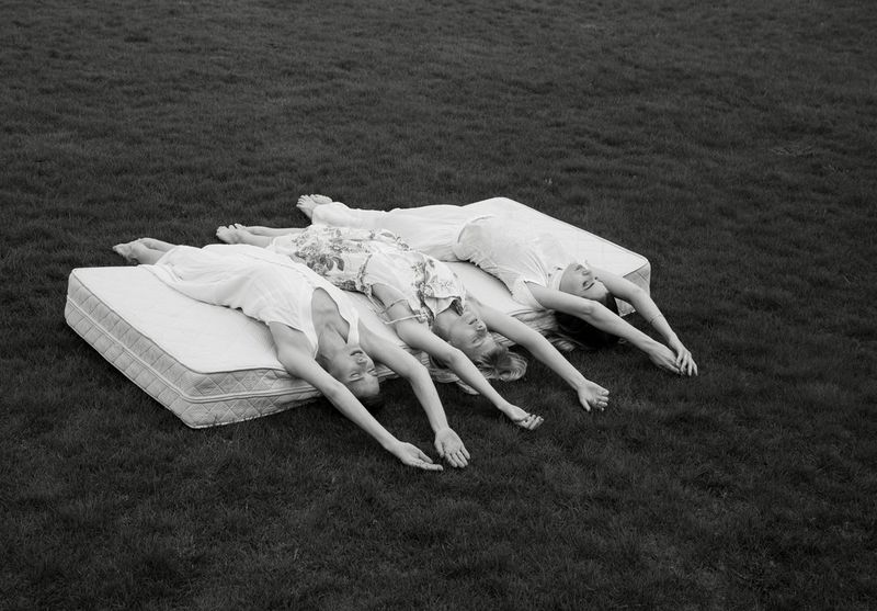 A black and white image of three sisters lying stretched out on a single mattress in a field.