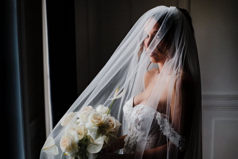 A bride in an embroidered, strapless dress, lit from the side, poses wearing a veil that covers her head and the bouquet of roses she is holding.