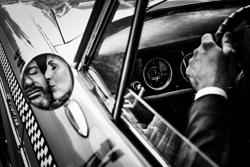 In this black-and-white wedding photograph, the wing mirror of a convertible reflects a woman kissing a man on the cheek. The man's left arm, dressed in a smart suit, can also be seen holding the steering wheel.