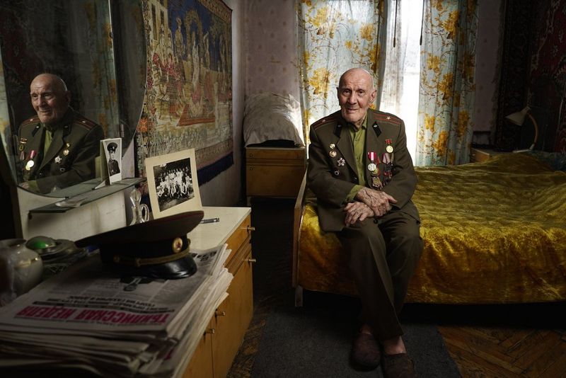An elderly Second World War veteran wearing his medals, seated on the end of his bed. His reflection is visible in the mirror on the wall.