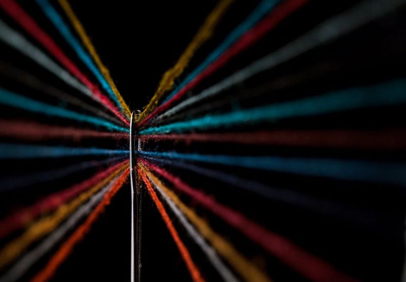 A close-up image, taken with a macro lens, of multiple multi-coloured fibres stretched through the eye of a needle.
