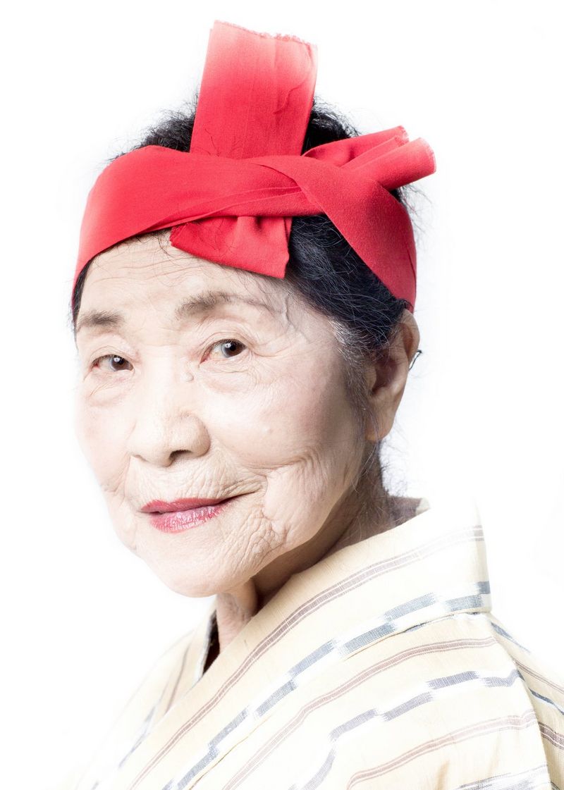 An older Japanese woman looks at the camera, wearing dark pink lipstick and with a red piece of fabric tied around her hair.