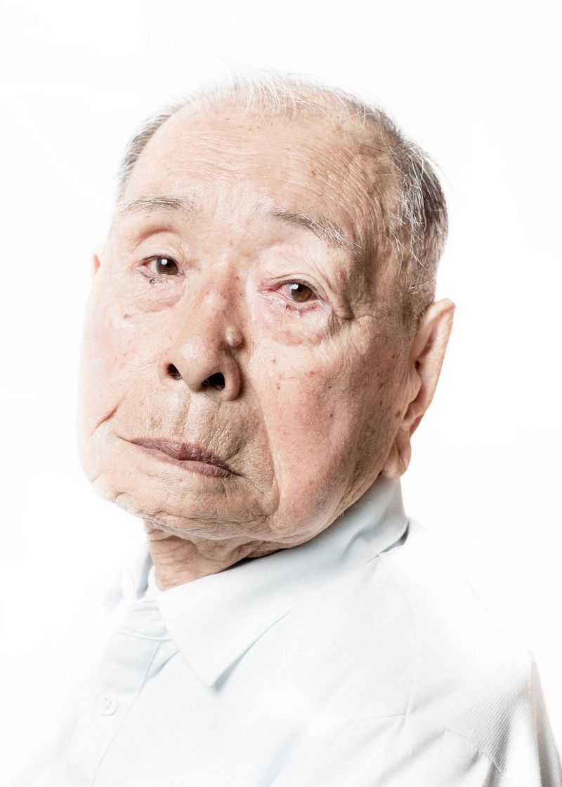An older Japanese man looks at the camera, wearing a white shirt in front of a white background.
