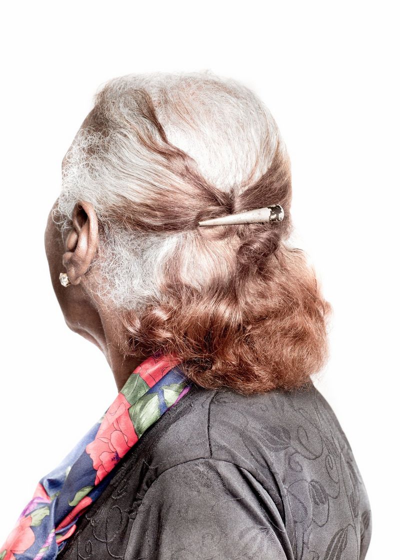 A woman is turned away from the camera, showing the back of her white and brown hair, which is held in a clip. She wears a grey top and a colourful scarf.