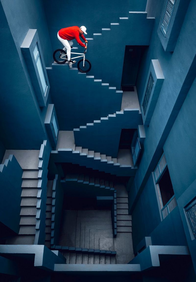 A BMX rider in a red top balances on the edge of a staircase in the blue stairwell of an apartment complex.