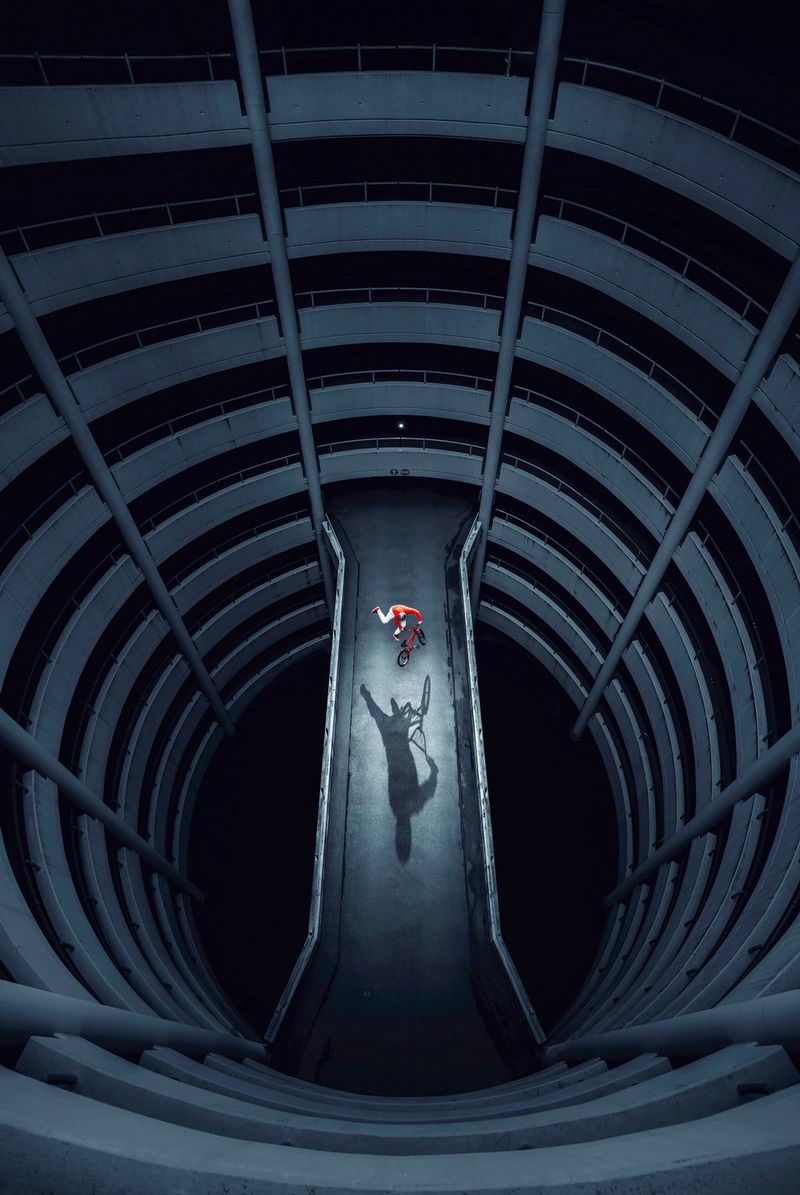 An overhead shot of a BMX rider performing a trick between levels in a dark, circular multi-storey car park.
