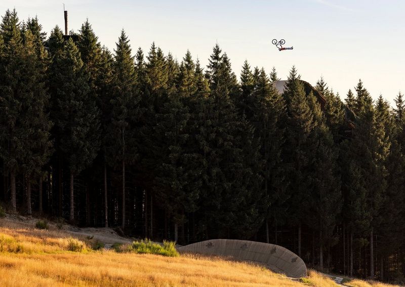 A BMX rider hanging from the handlebars of his bike as he soars through the air above a line of conifers on a sloping course.