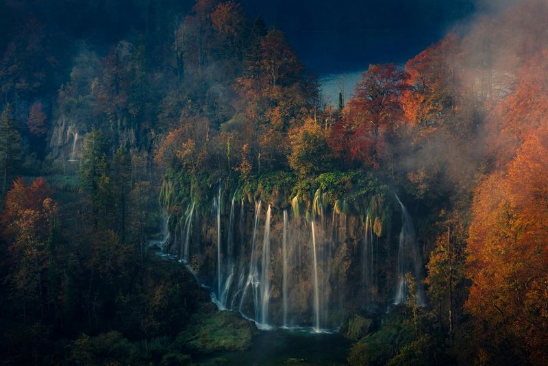 A landscape shot of a waterfall flowing down a cliff surrounded by autumnal trees, photographed with a long exposure by Canon Ambassador Luka Vunduk.