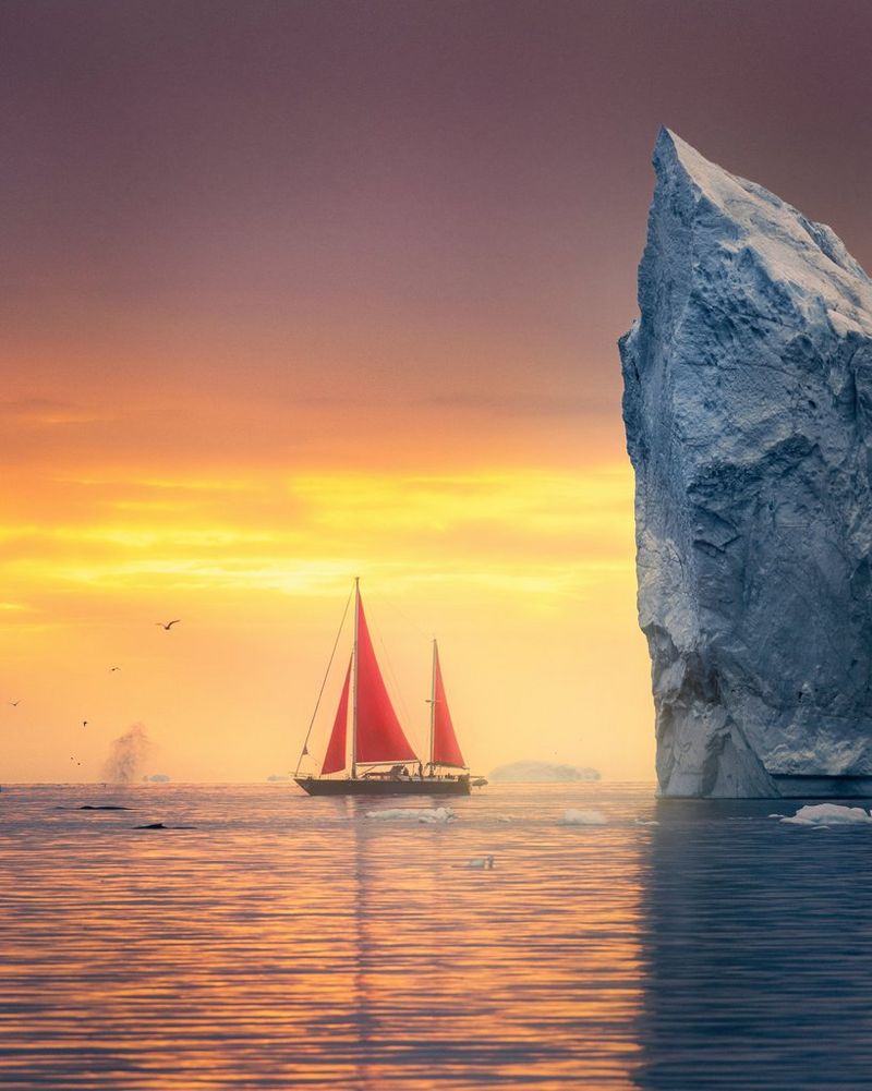 A boat with red sails photographed next to an iceberg under an orange sky in Greenland. Spray from two whales can be seen to the left of the boat. Taken on a Canon EOS R5 by Canon Ambassador Luka Vunduk.