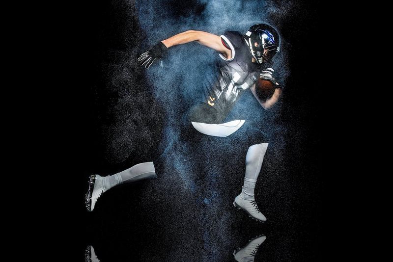 An American football player poses mid-run, clutching a ball in front of him with one arm and leg outstretched behind, on a black background.