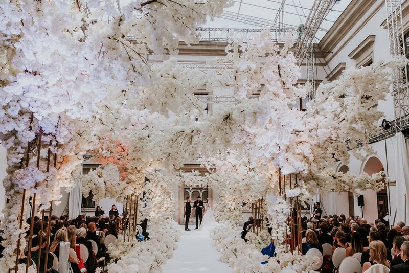 Un couple échange ses vœux au château Ujazdów de Varsovie sous le regard de ses nombreux invités. À la fois imposante et somptueuse, la pièce est remplie de fleurs blanches, du sol au plafond. Photo prise par Malachite Meadow avec un Canon EOS R6.