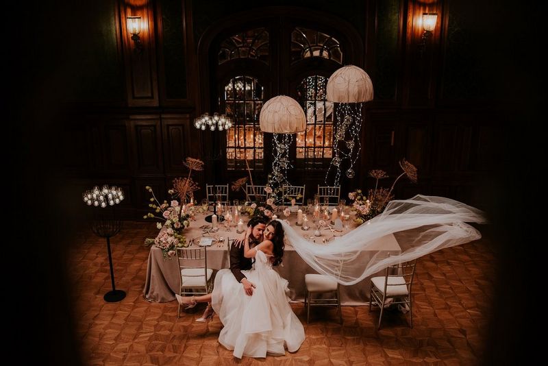 Photo prise du dessus sur laquelle on a ajouté une vignette noire. Deux mariés assis s'embrassent amoureusement près d'une table décorée avec soin. Photo prise par Malachite Meadow avec un Canon EOS R6 Mark II.