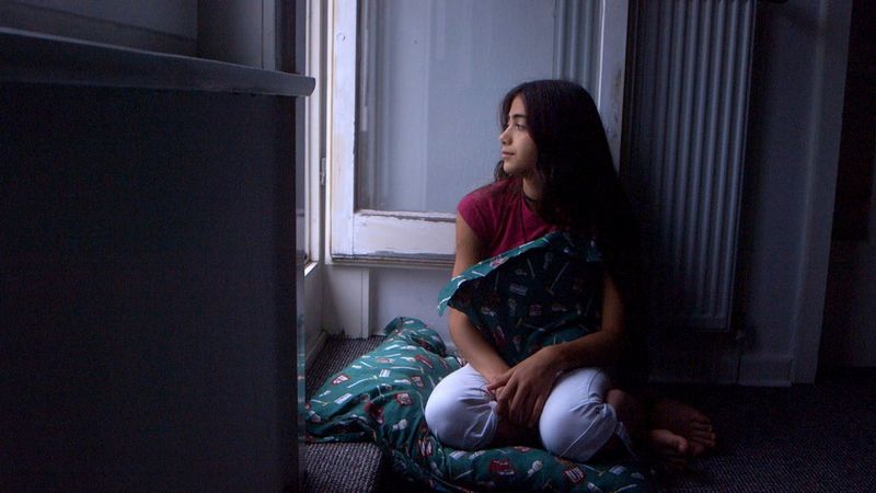 A girl kneels by an open window looking out, holding a patterned pillow.