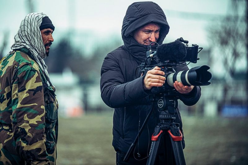 A man in a thick black coat with the hood up stands beside a Canon cinema camera, adjusting the lens. Next to him stands a man in a camouflage jacket and headscarf, looking at the camera.