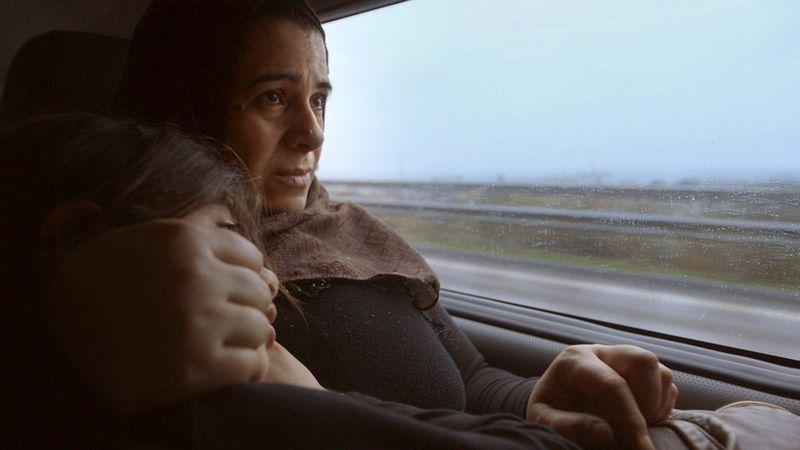 A woman cradling her young child as they sit in the back of a car, a rainy motorway moving past out of the window.
