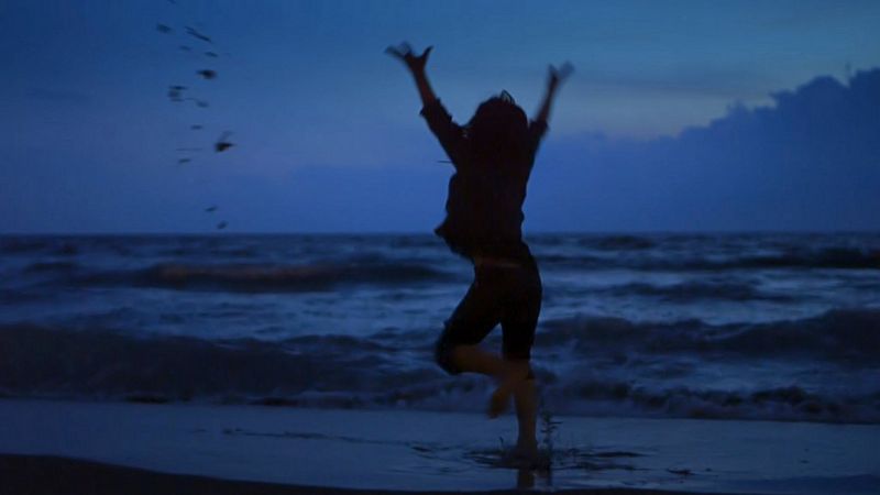 A woman is silhouetted dancing on the beach after sunset, her arms raised in the air above her head.