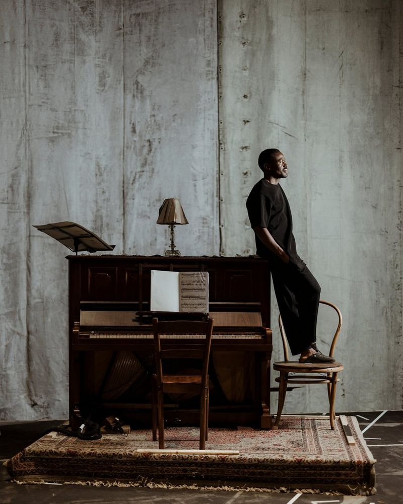 A portrait of a man, in side profile, standing on a chair and leaning against a piano.