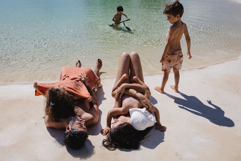 A couple lying on a beach with their four children – triplets and a baby. They are holding the baby and one of the triplets, while the other two children play in the sea.