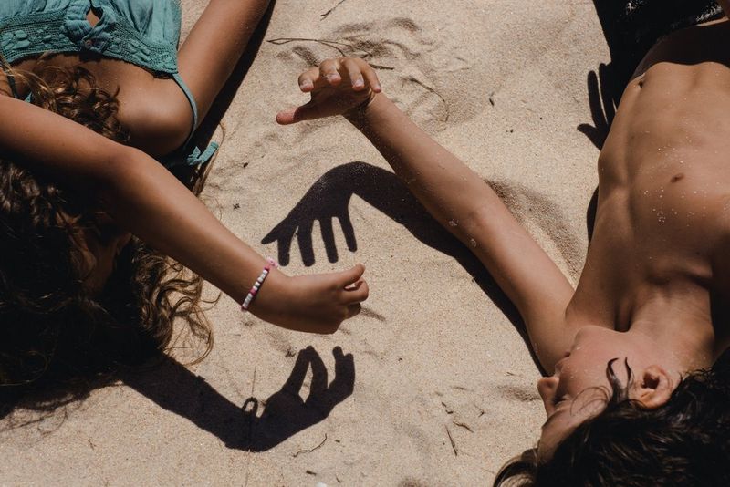Two children, who are lying down on their backs, make hand shadows in the sand on a beach in Portugal. Taken on a Canon EOS R by Marisa Martins.
