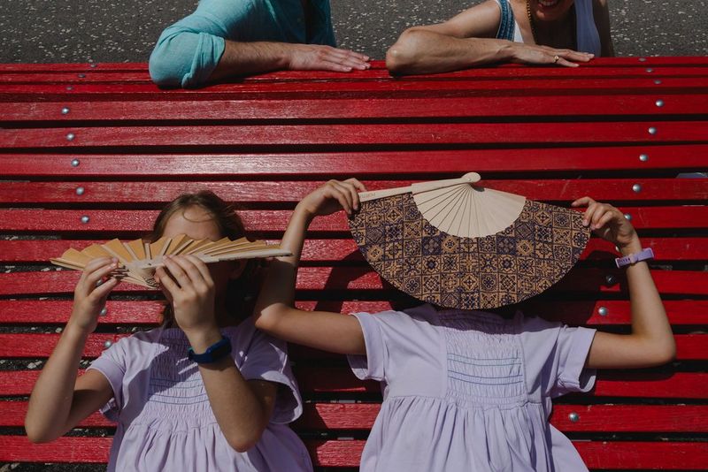 Two young girls in matching lilac dresses lie back on a red bench, their faces obscured by fans.
