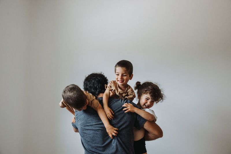 A father with his back turned to the camera holds three small children at once. The children are laughing and attempting to clamber over his shoulder. Taken on a Canon EOS R5 by Marisa Martins.