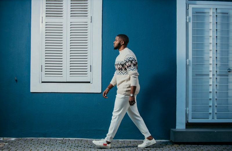 A man in a patterned jumper, white trousers and white shoes walks near a blue wall with white windows. Taken on a Canon EOS R by Canon Ambassador Maropeng Vushangwe.