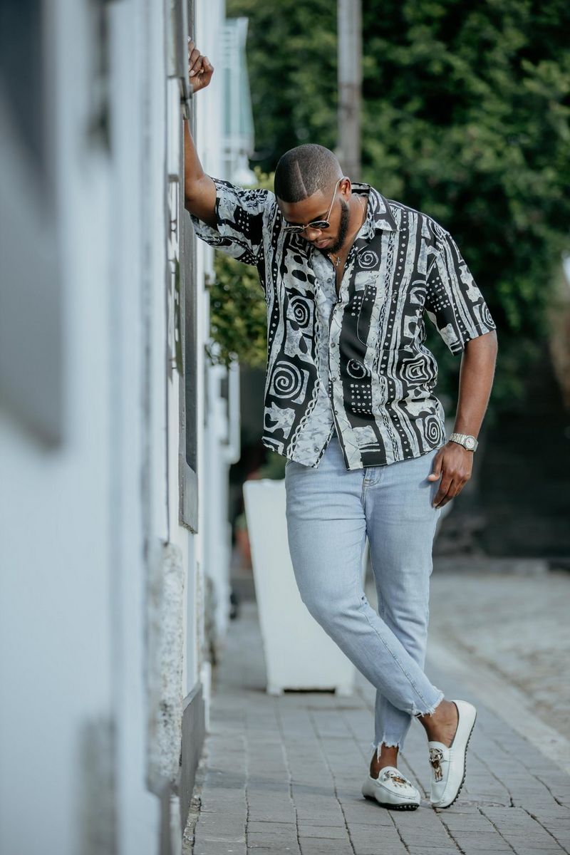 A man in a black and white patterned shirt leans against a wall and looks down at the ground, in a photo taken on a Canon EOS R by Canon Ambassador Maropeng Vushangwe.