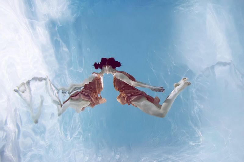 An underwater portrait of a woman floating with just her head above the surface, her softly distorted reflection mirrored in the water.
