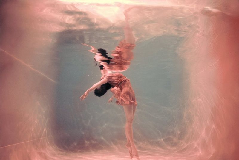 An underwater portrait of a woman with her arms thrown back behind her head, her distorted reflection visible in the water above.