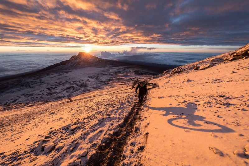 Three men walk on a snow-covered mountain while carrying bicycles as the sun peeks through the clouds.