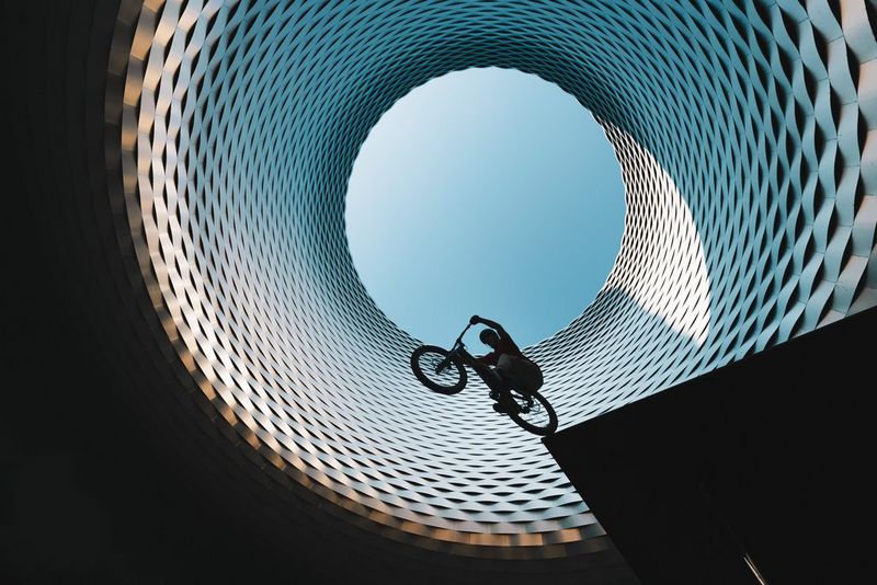 A mountain biker balances in silhouette on the roof of a bus station, framed by the circular opening in the roof of Messe Basel.