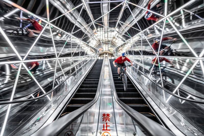 A mountain bike rider descends an escalator in a modern steel and mirrored structure.