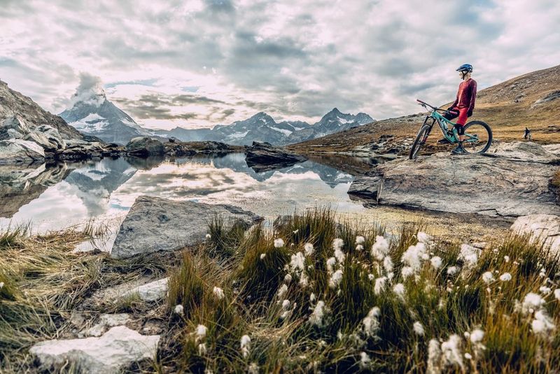 A cyclist stands aboard their bike at the edge of a lake with mountain scenery in the background. 