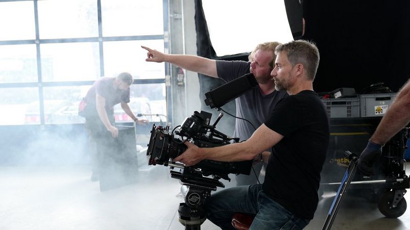 A man tilts a Canon camera upwards on a dusty film set, while a second man points out something in the distance to him.