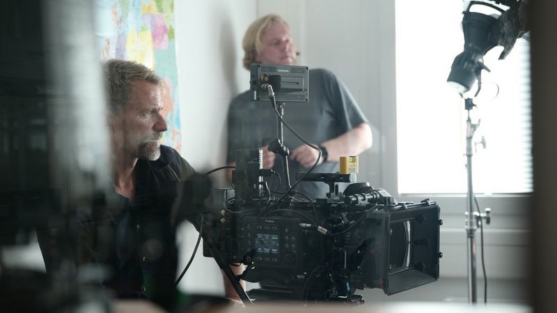 A man sits in a room behind behind a large Canon cinema camera. Another man is stood in the corner of the room.