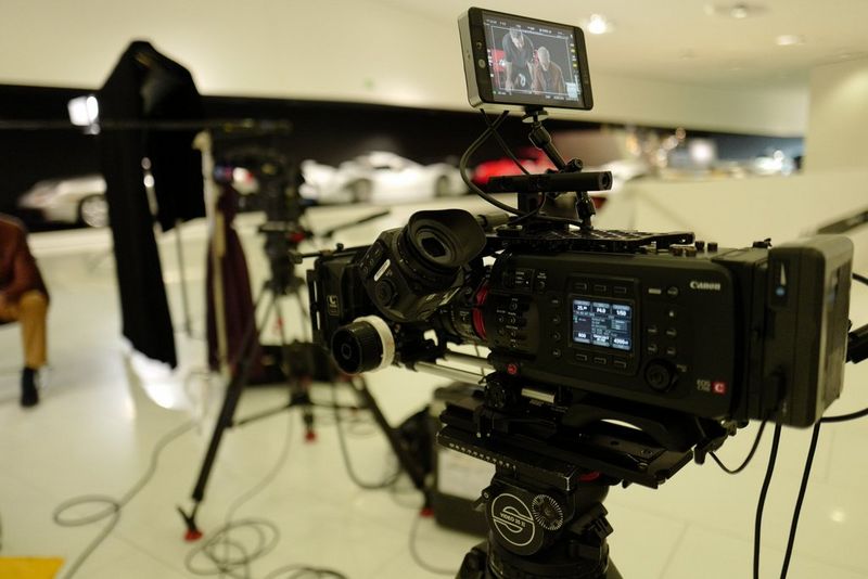 A close-up of a Canon EOS C700 camera, which is positioned on a tripod in a large garage filled with gleaming racing cars.