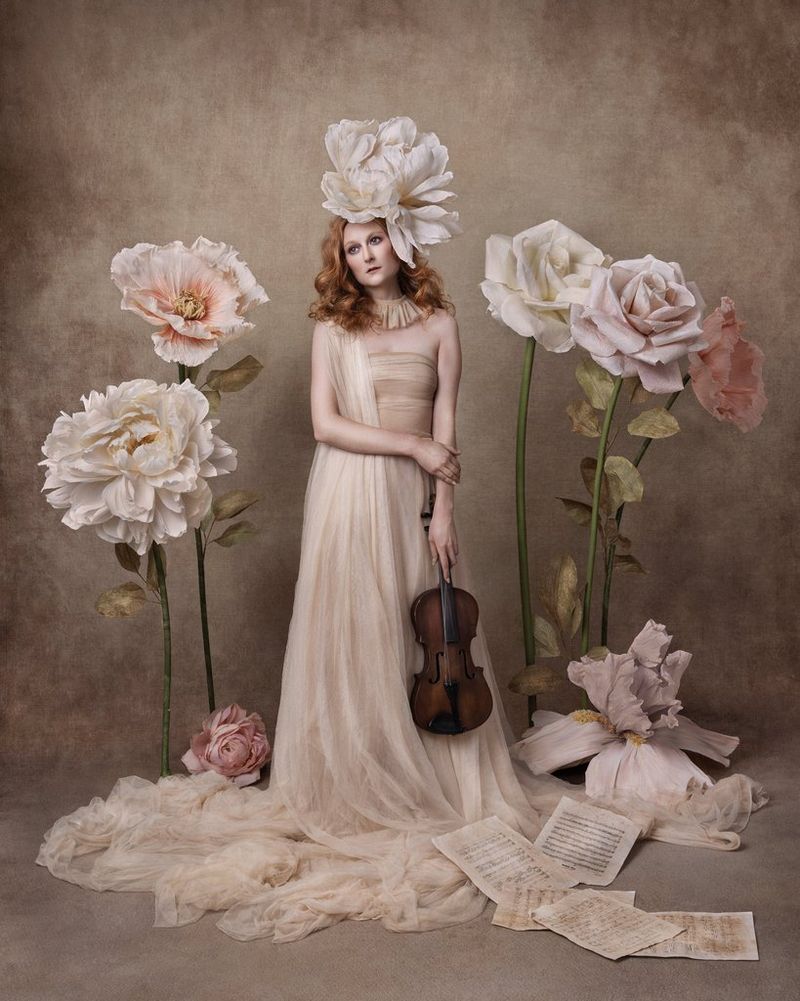 A painterly-style portrait of a woman, holding a violin and with a large floral headpiece, standing in the middle of a group of oversized flowers.