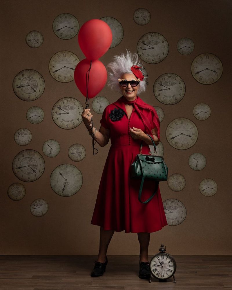 A portrait of an older woman with white spiky hair, wearing a bright red dress and oversized sunglasses and holding two red balloons, against a backdrop of clock faces. Taken on a Canon EOS R5 by Martina Wärenfeldt.