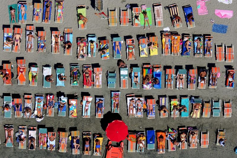 Photographed from above, rows of sunbathers enjoy a day at the beach in Ostia, Italy. A single red umbrella is the only form of shade. 