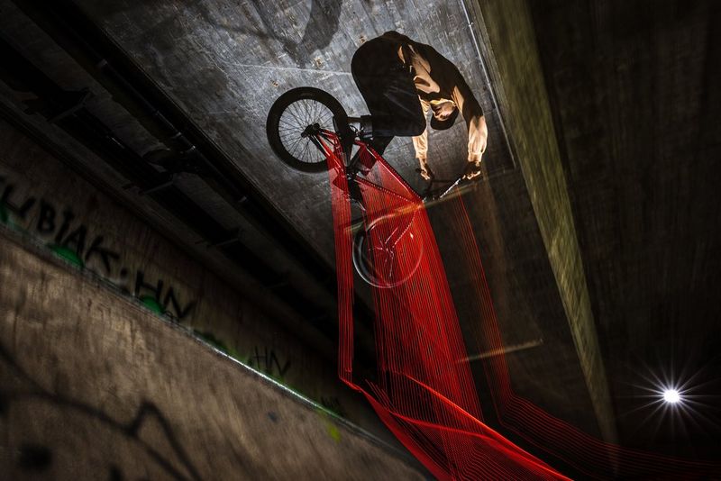 Des lumières rouges montées sur le cadre d'un vélo créent des traînées lumineuses, sur une image en pose longue du pilote de BMX Miha Glavič qui effectue des cascades sous un pont. Photo prise par l'ambassadeur Canon Matic Borković, avec un Canon EOS R7.