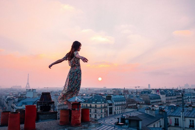 A woman in a long floaty dress stands on a rooftop overlooking the streets of Paris. Her arms are stretched out behind her.
