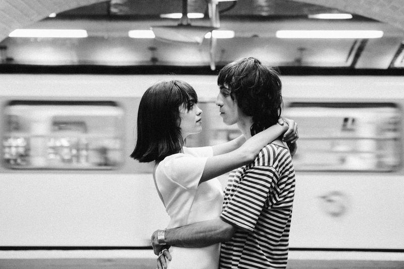 A black and white image of a woman and man locked in an embrace on a Paris Métro platform. Behind them, a train speeds past.