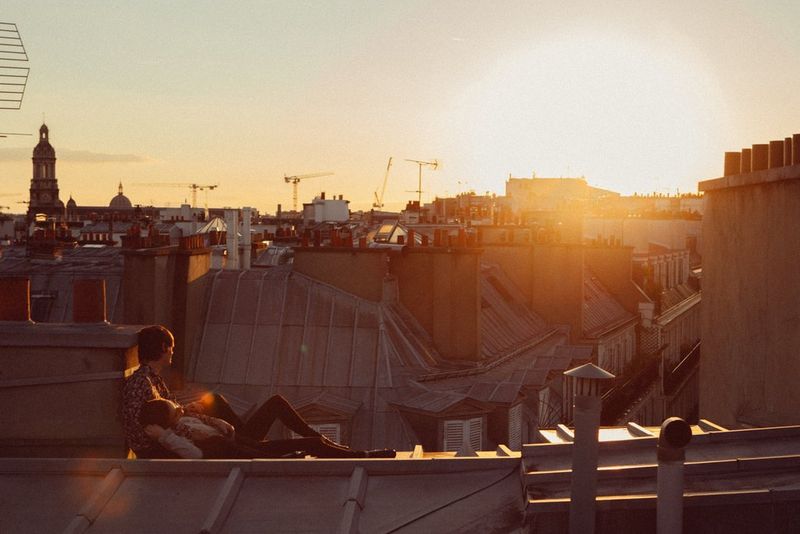 A woman lies between the legs of a man who is sat on a rooftop overlooking a city skyline.