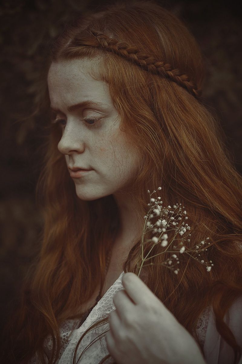 A side portrait of a woman with braided red hair staring downwards in a melancholy manner.